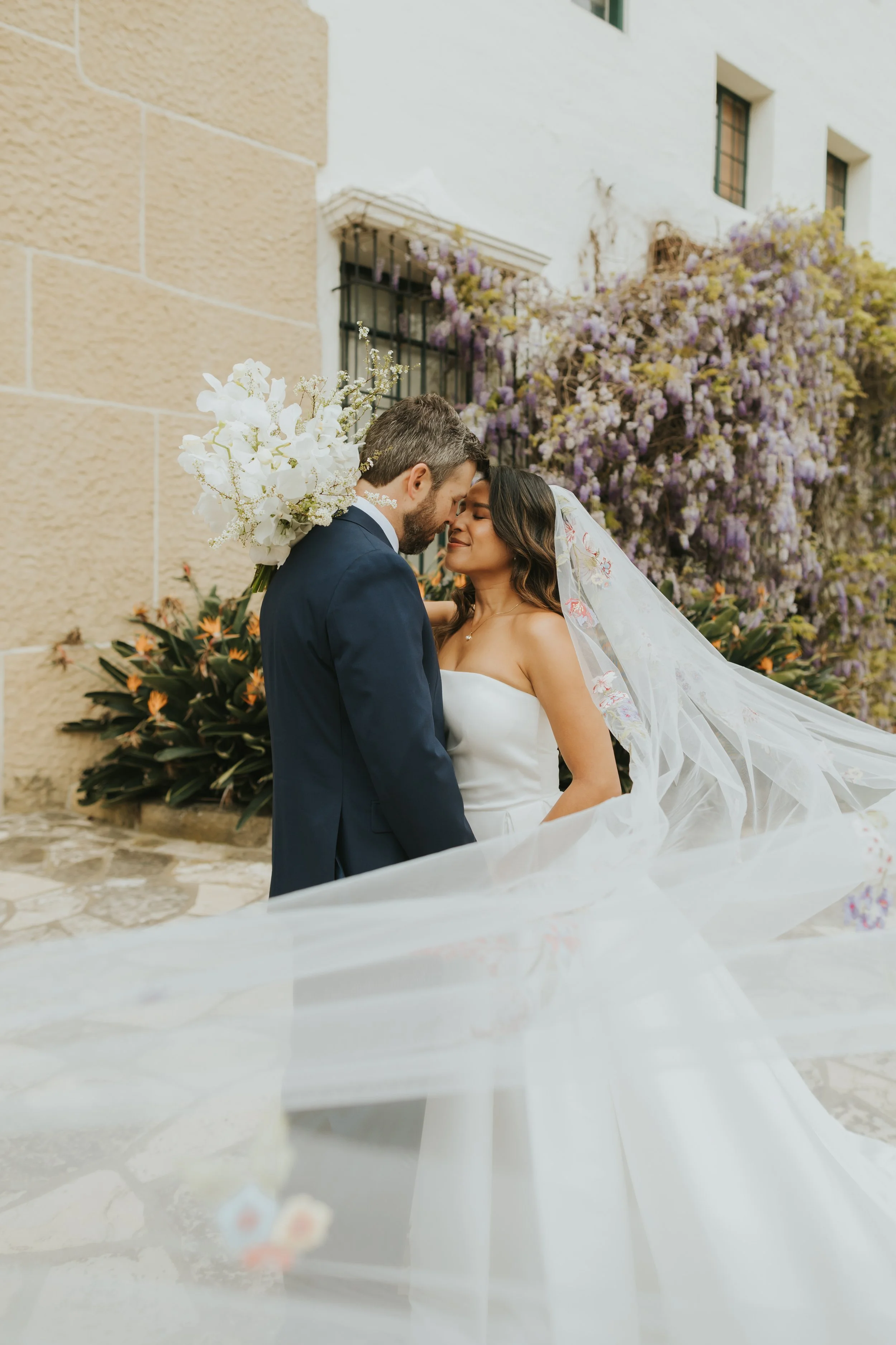 A bride and groom sharing a tender moment outside a building with blooming purple and white flowers in the background.
