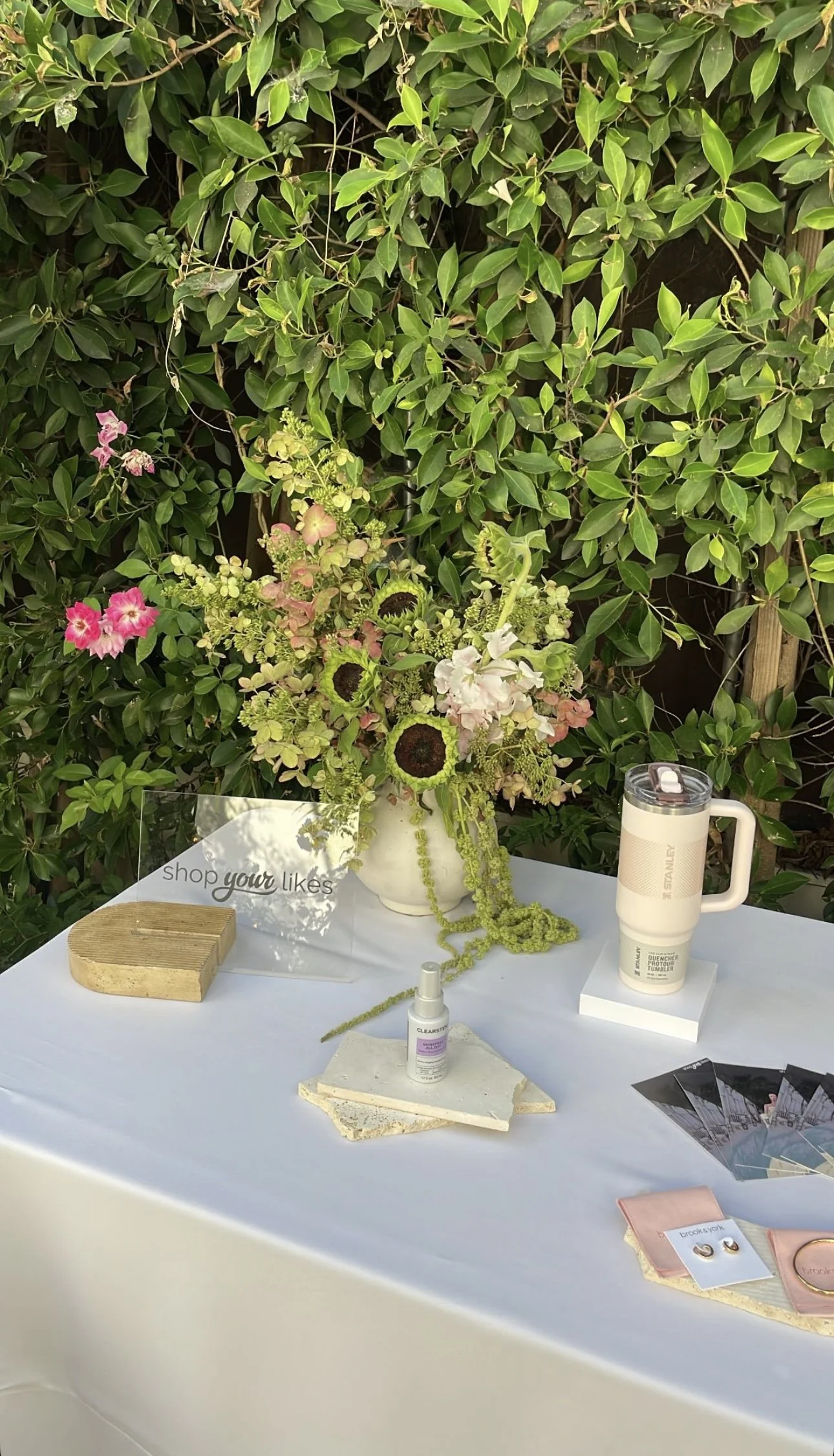 A display table with jewelry and skincare products, a floral arrangement with pink, white, and brown flowers, and a backdrop of green leafy bushes.