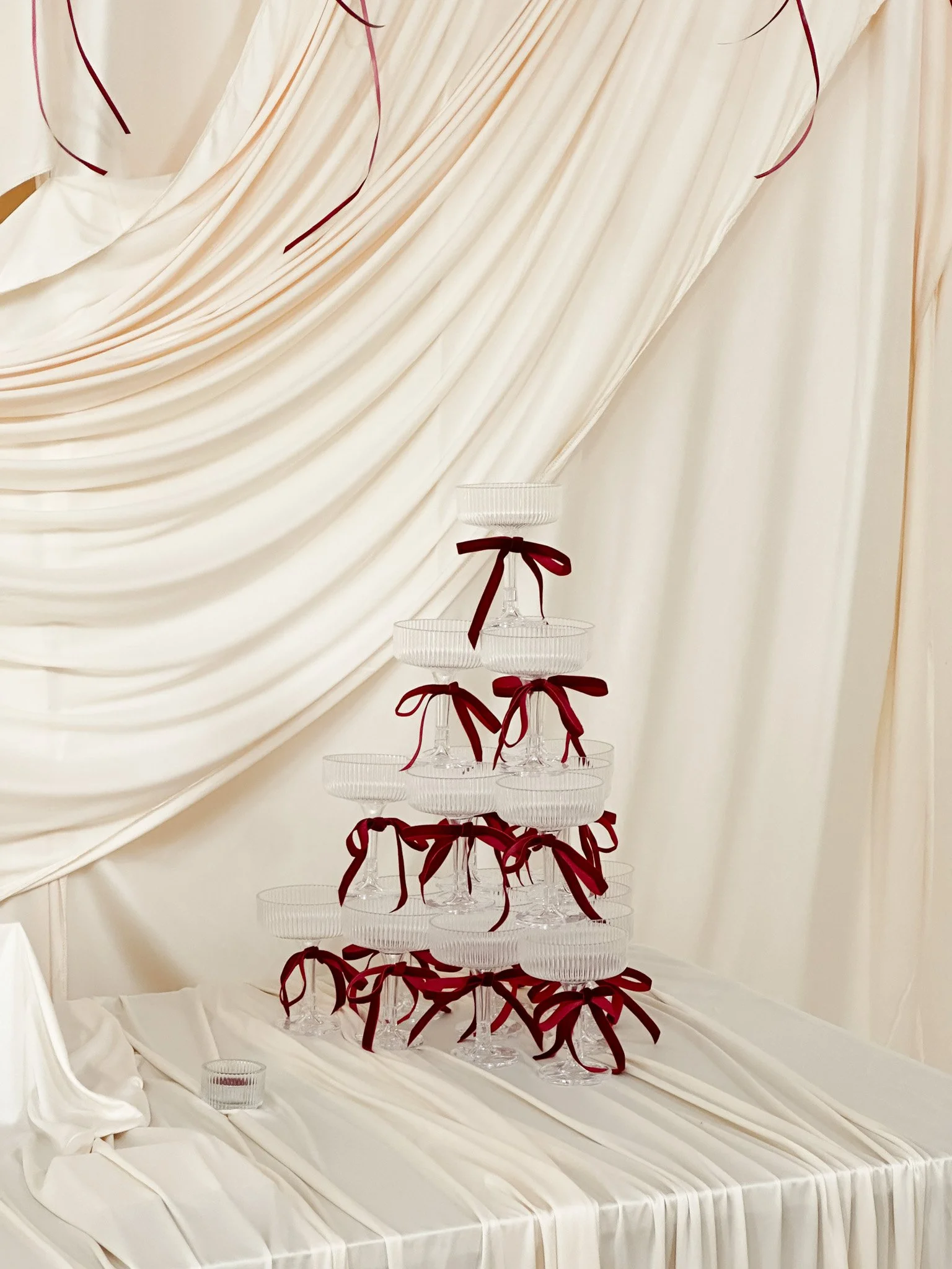 A multi-tiered glass dessert stand with red ribbons tied around each layer, set against a backdrop of cream-colored draped fabric.