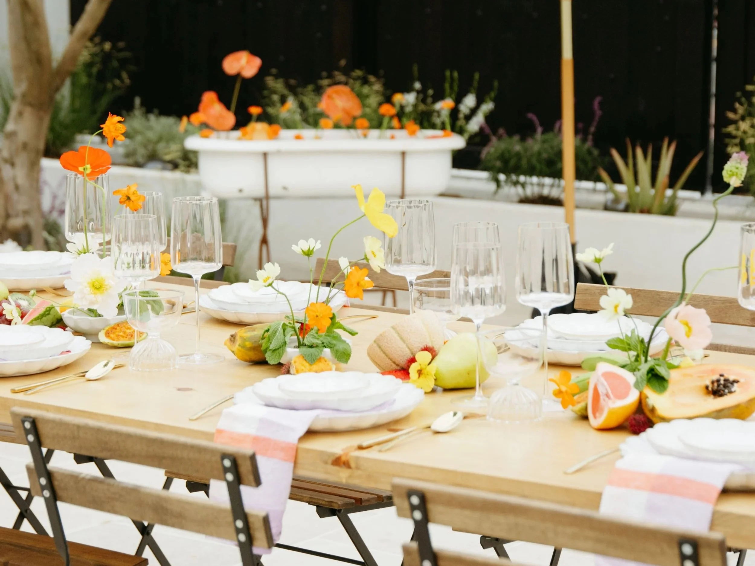 A wooden dining table set for a meal with white plates, clear glasses, and colorful flowers as centerpieces in a bright outdoor setting with a garden background.