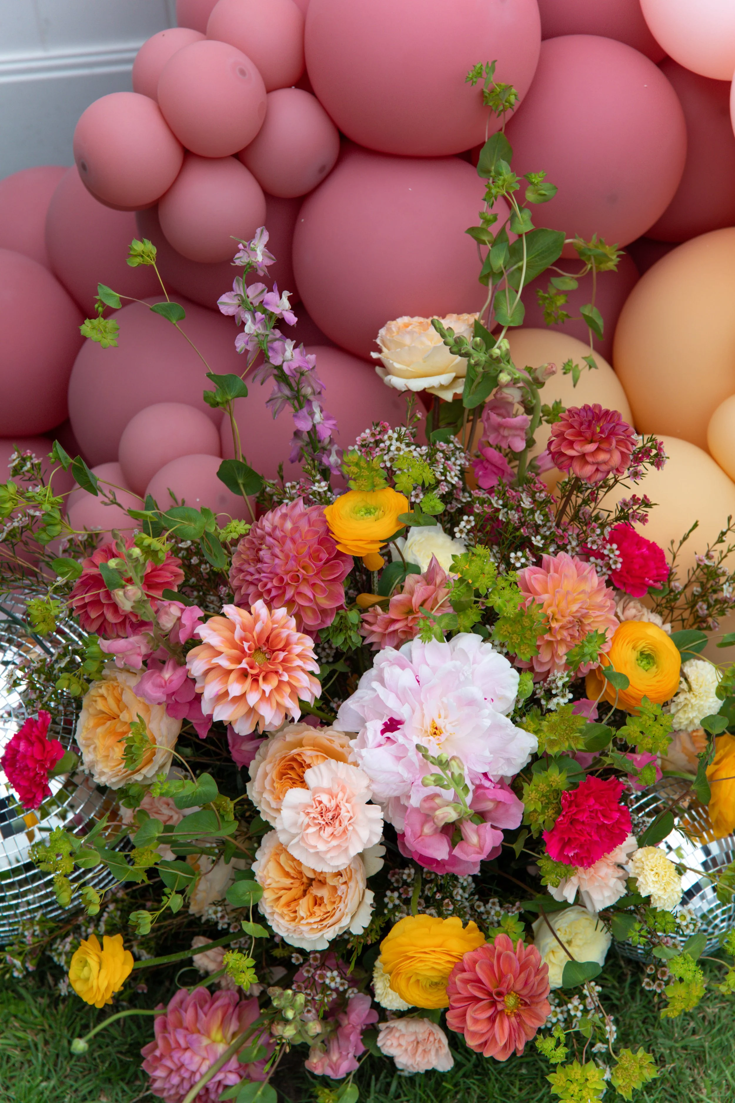 A colorful bouquet of various flowers including pink, yellow, orange, and white blooms, with green leaves, placed in front of a backdrop of pink and peach balloons.