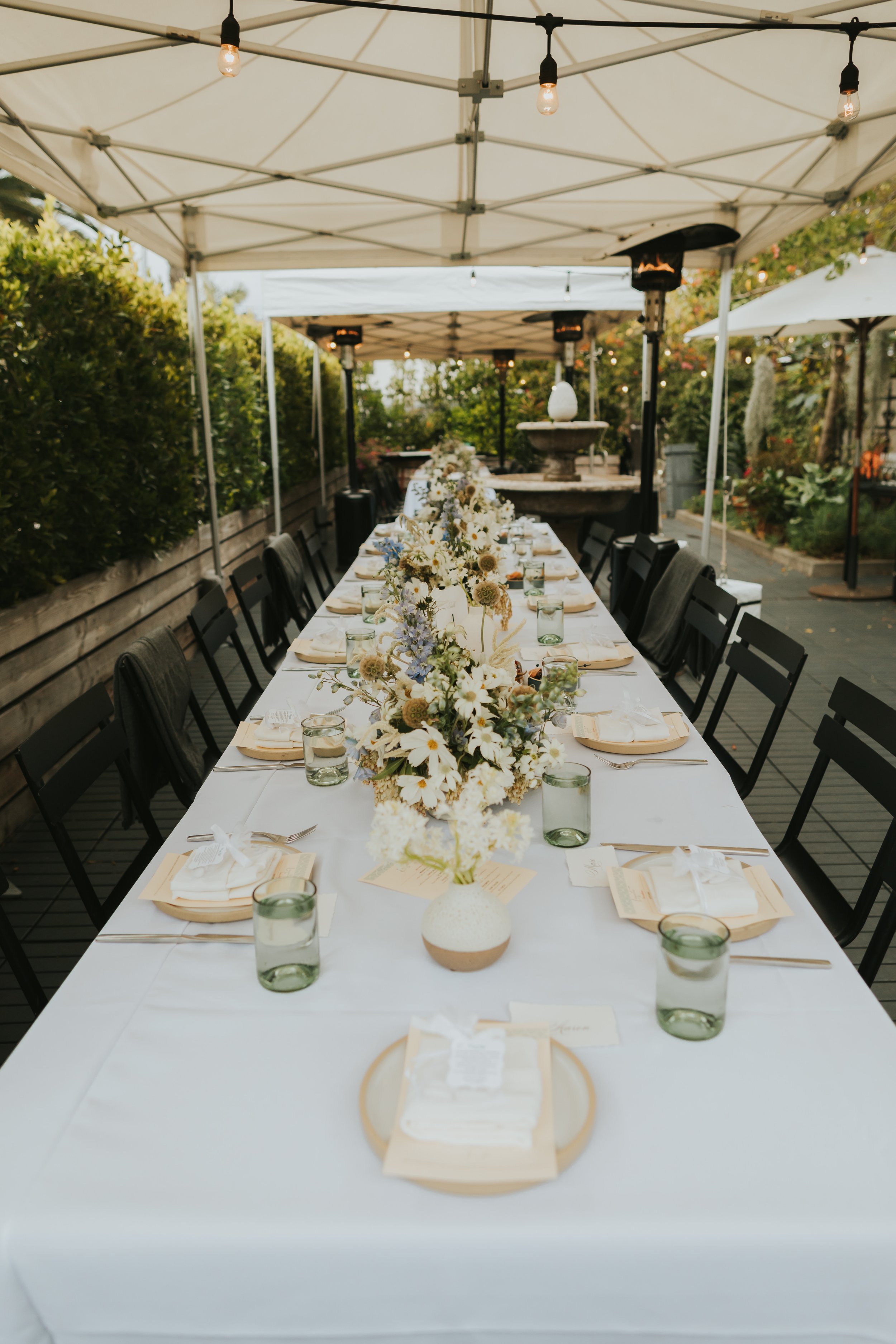 A long outdoor dining table set under a canopy with white tablecloths, floral centerpieces, plates, napkins, glasses, and utensils, surrounded by black chairs, with string lights overhead and a garden in the background.