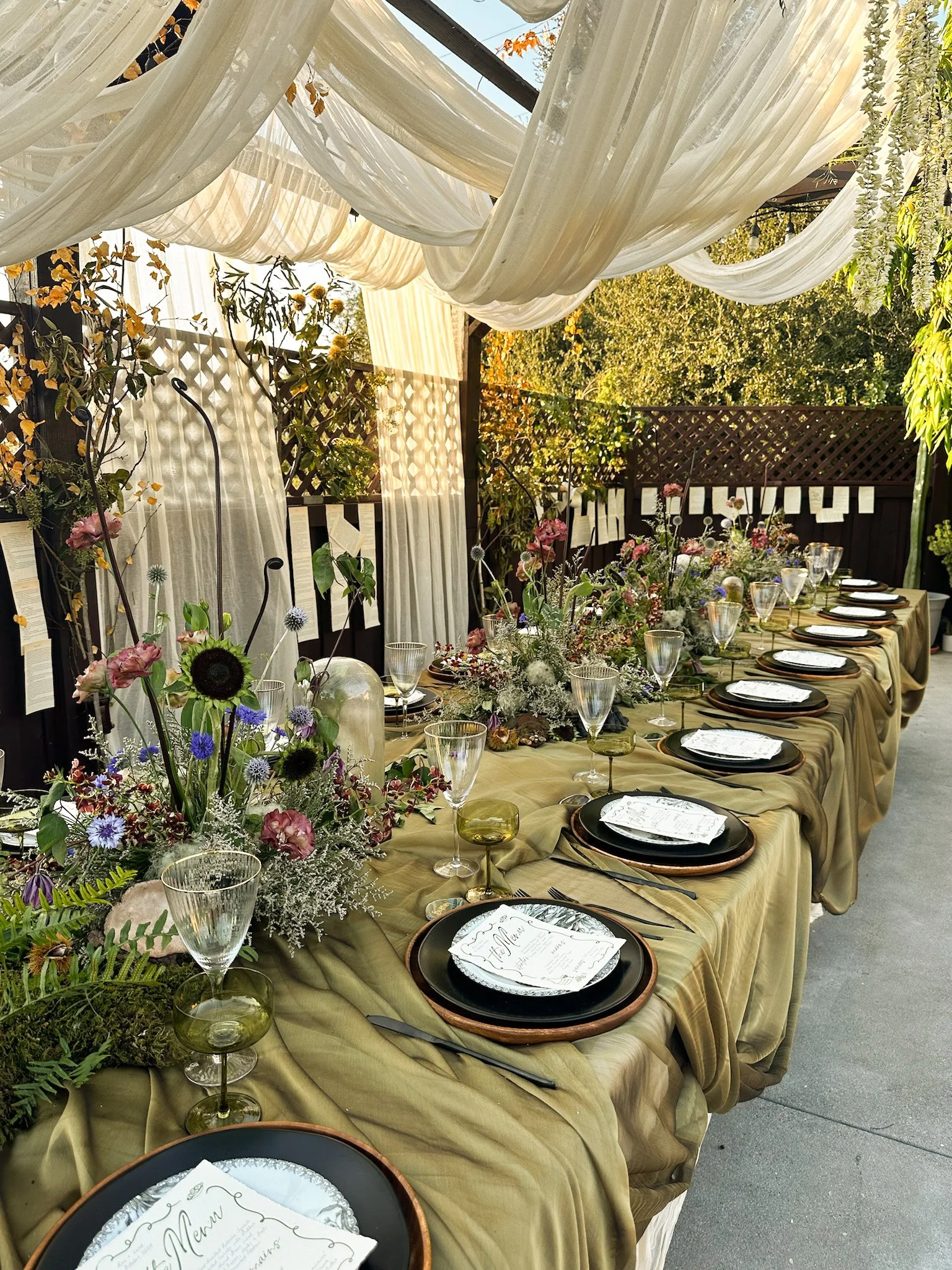A beautifully decorated outdoor dining table with a beige tablecloth, floral centerpieces, black plates, and clear glassware under a draped fabric canopy surrounded by trees and greenery.