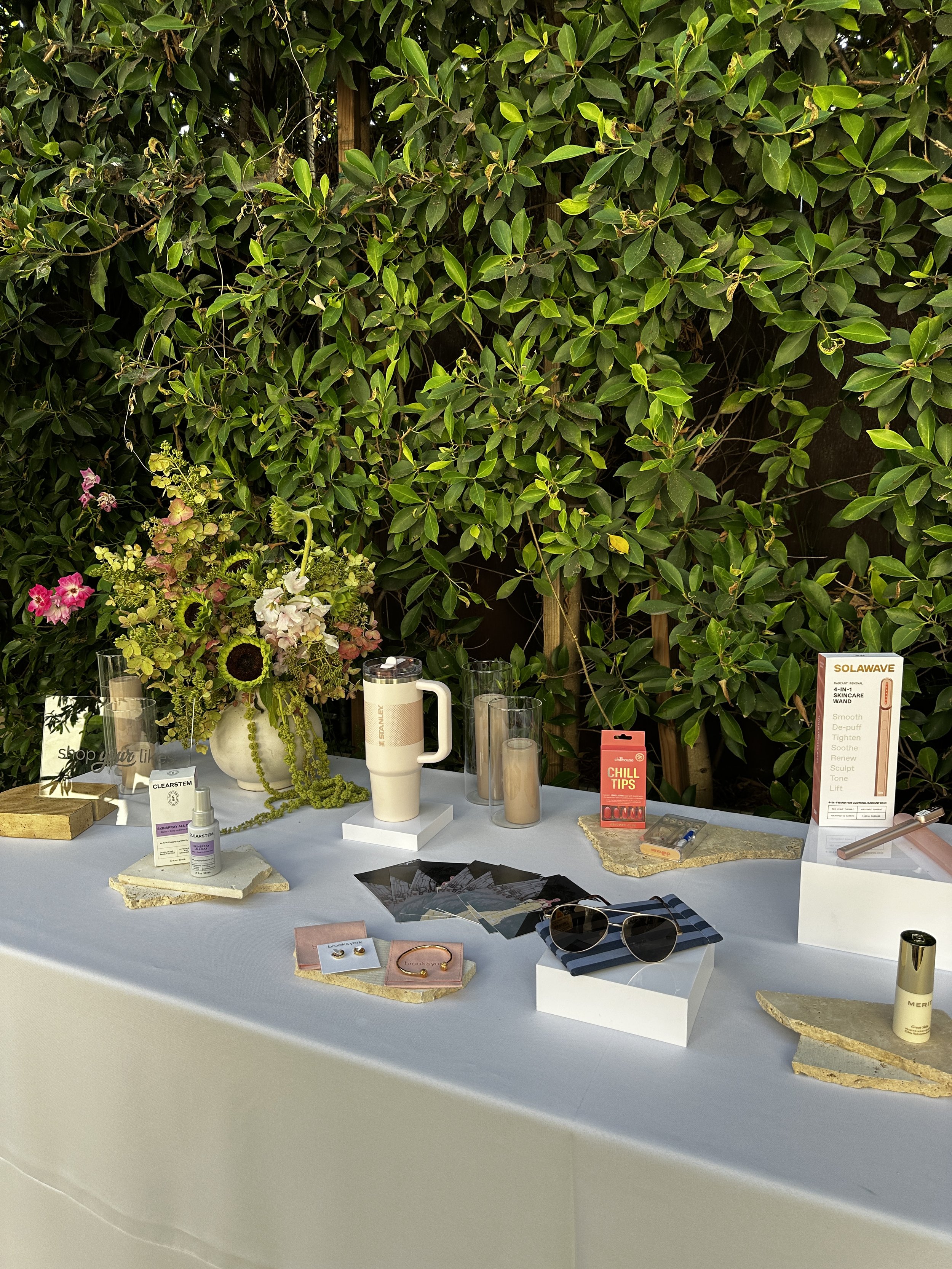 Brand Display table with skincare, sunglasses, jewelry, and beauty products in front of a lush green hedge background.