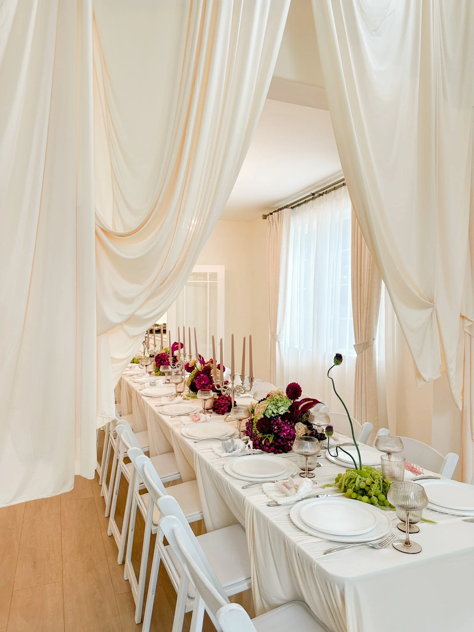 Elegant dining room set for a special occasion with a long table decorated with floral arrangements, candlesticks, and glassware, framed by white curtains and draped fabric overhead.