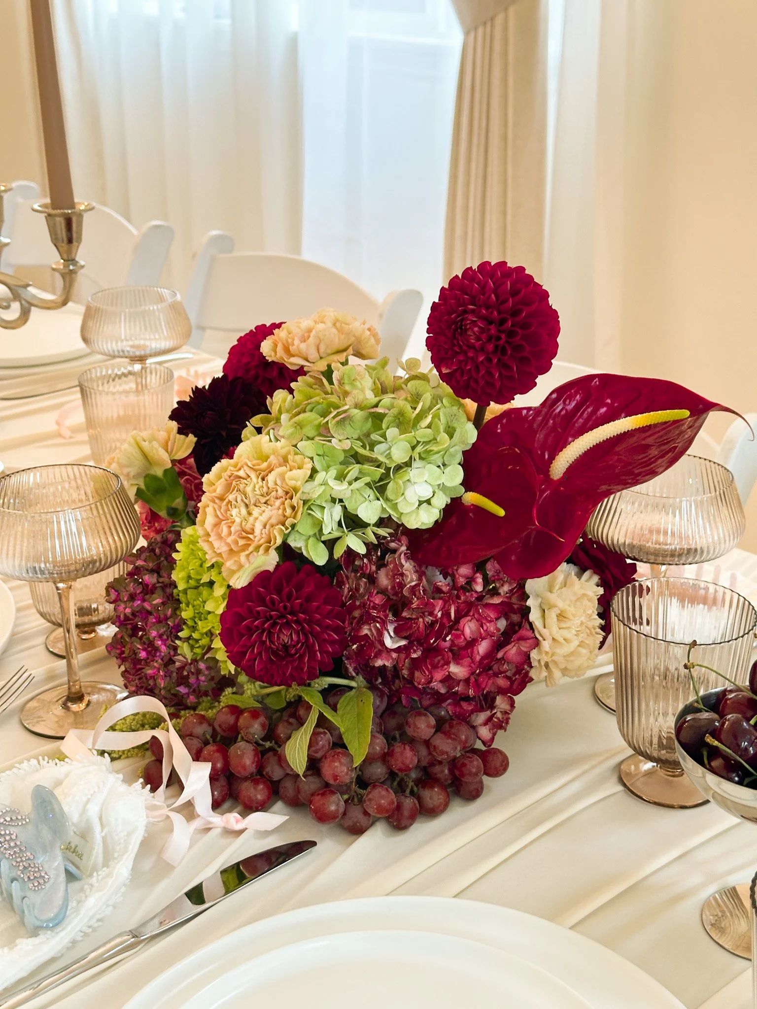 A table centerpiece featuring a large floral arrangement with red, cream, and light green hydrangeas, and red calla lilies, surrounded by glass candle holders and grapes at an elegant dinner setting.