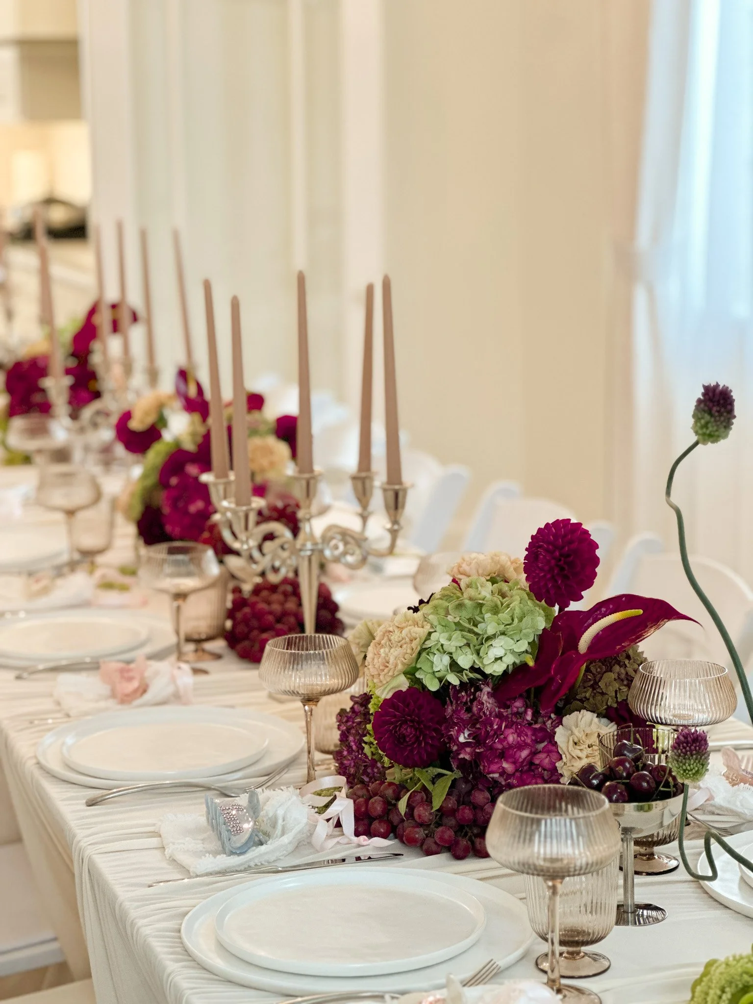 Elegant all white table setting with a centerpiece of purple, pink, and white flowers, silver candlesticks, and glassware, arranged on a white tablecloth at a bridal shower event.