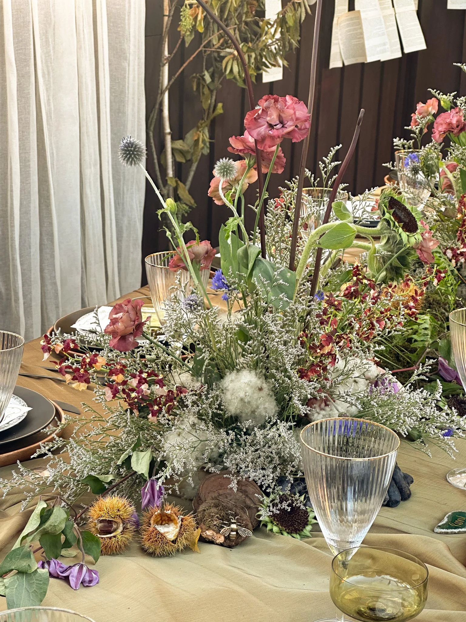 A floral centerpiece on a dining table featuring pink, white, and purple flowers, with green leaves and decorative elements, surrounded by glassware and table settings.