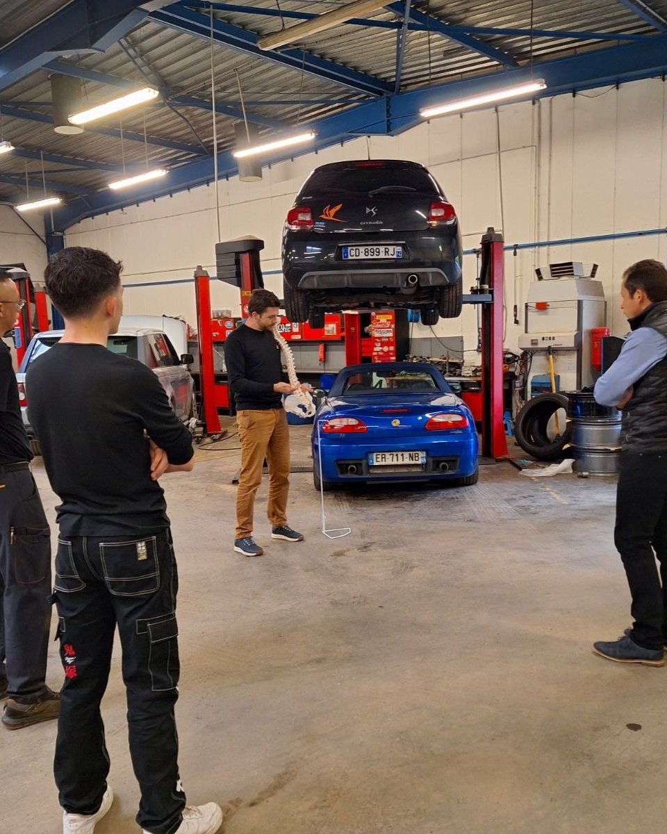 Un groupe de personnes observant une démonstration dans un atelier de réparation automobile. Un modèle de voiture noire est sur un pont élévateur, une voiture bleue est stationnée en dessous. La personne au centre tient un outil de réparation ou de diagnostic.