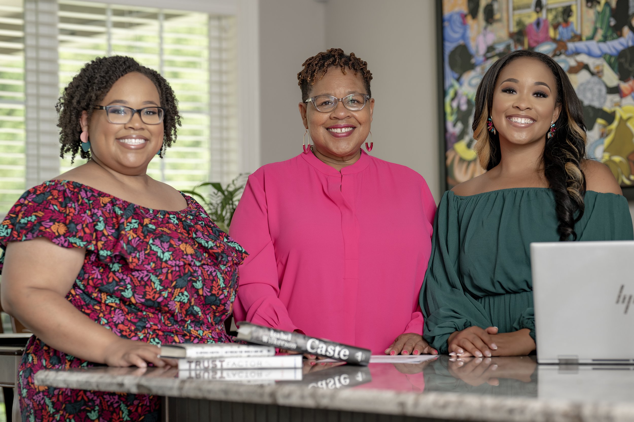 Three women standing together in an office space, smiling at the camera. The woman on the left has glasses and short curly hair, wearing a colorful floral dress. The woman in the middle has glasses and short hair, wearing a bright pink dress. The woman on the right has long wavy hair, wearing a dark green dress, and colorful earrings. There are books and a computer on the countertop in front of them.