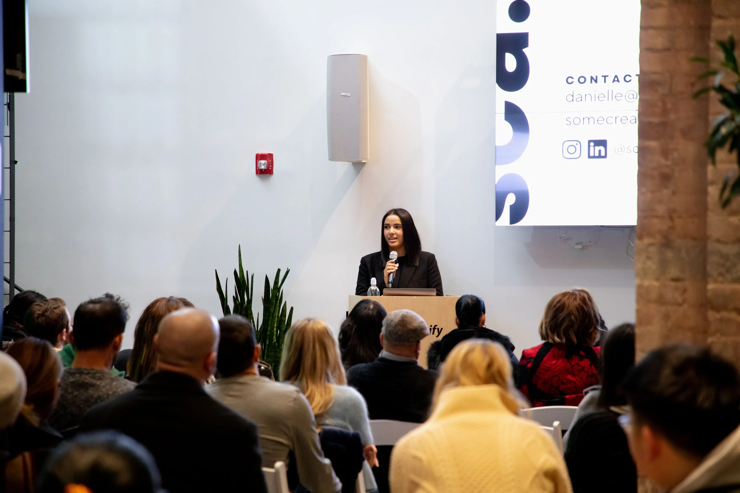 A woman giving a presentation at a conference, standing behind a podium with a laptop, microphone, and water bottle, while an audience listens in a modern indoor venue with white walls, a large screen displaying contact information, and a plant.