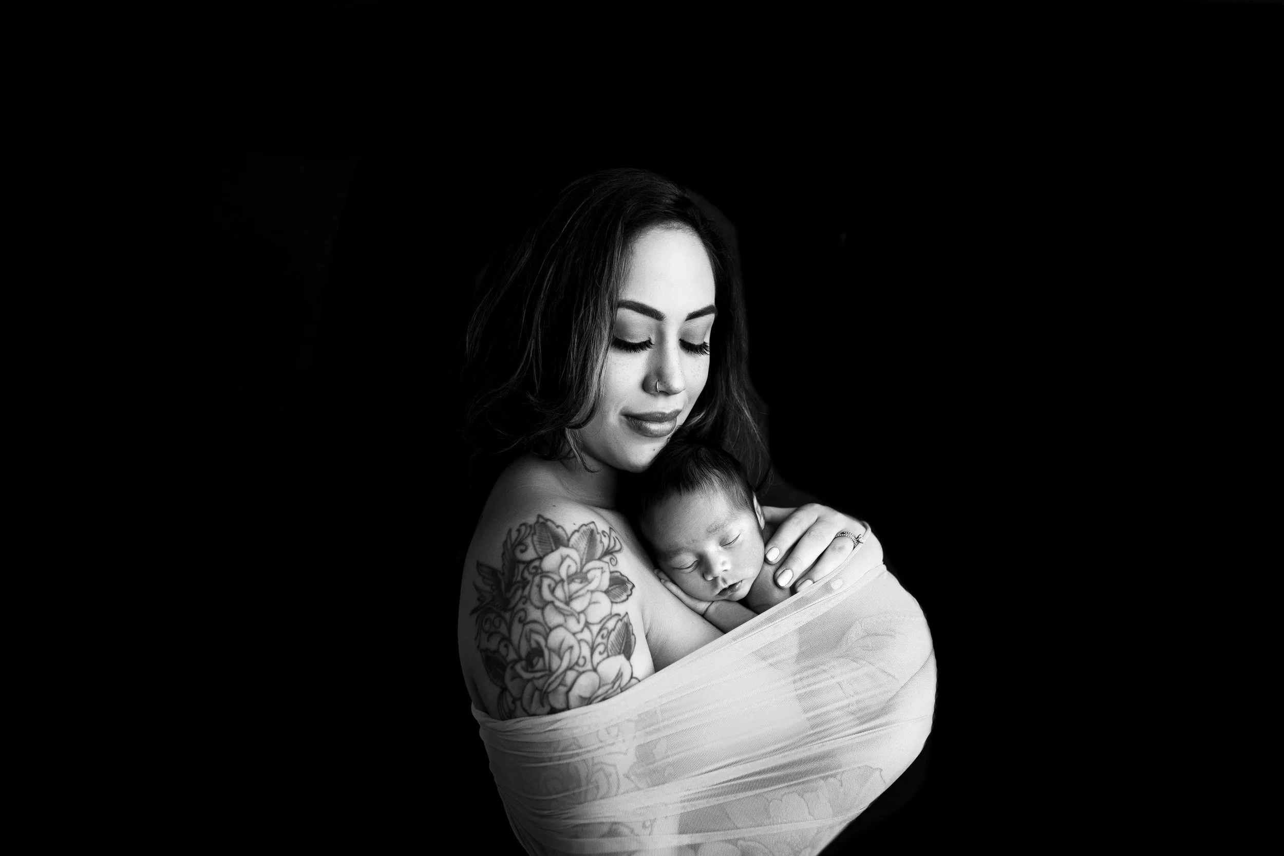 Emotional black and white portrait of a mother with a floral shoulder tattoo, cradling her sleeping newborn baby wrapped in a sheer fabric, with her eyes closed in contentment against a dark background.