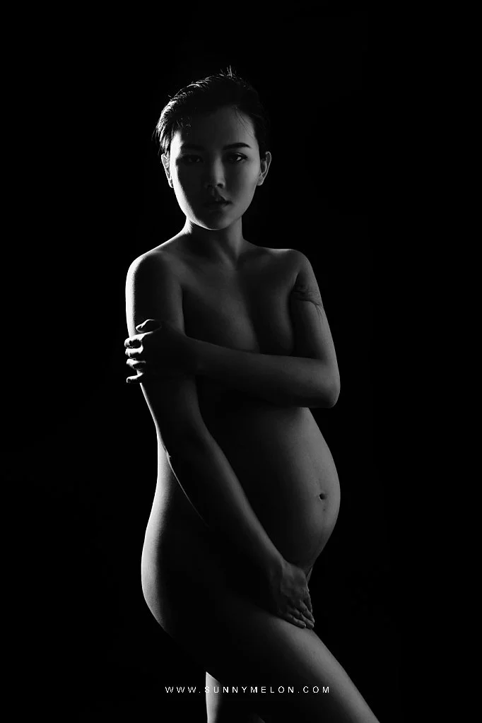 Sunnymelon Photography powerful black and white fine art maternity portrait. The expectant mother with short hair is posed with her arms crossed, dramatically lit from the front against a deep black background, highlighting her pregnant belly and str