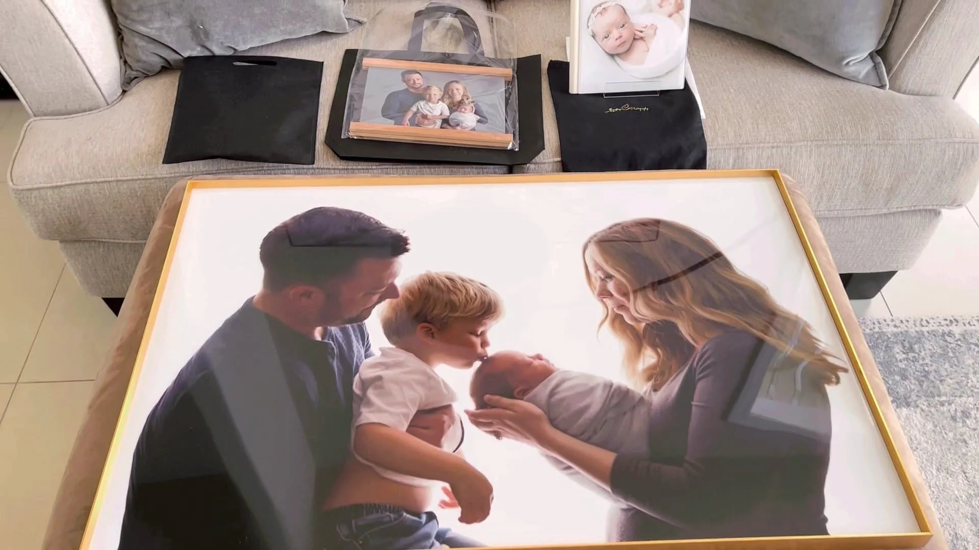 A collection of custom photography products displayed on an ottoman and sofa, featuring a large gold-framed family portrait, a smaller wood-trimmed frame, and a leather newborn photo album.