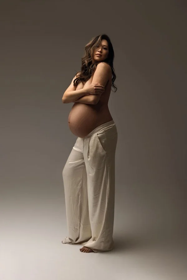 Sunnymelon Photography bold modern maternity portrait of an expectant mother. She is posing topless with arms crossed, wearing flowy white trousers that highlight her pregnant belly in a studio setting.