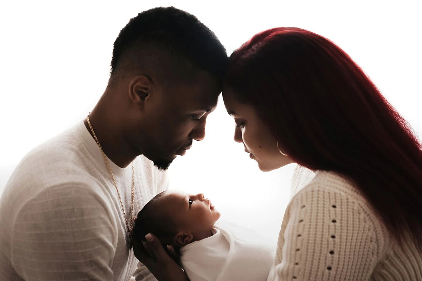 Backlit portrait of parents with foreheads touching, lovingly gazing down at their newborn baby against a bright white studio background.