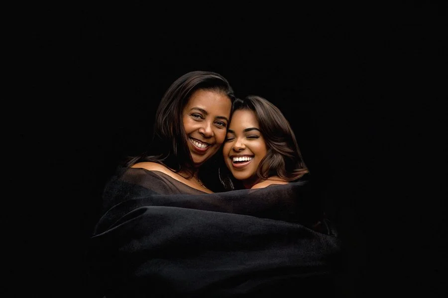 A high-end black and white style portrait of a mother and daughter laughing together, wrapped in a luxurious black fabric drape against a solid dark background.