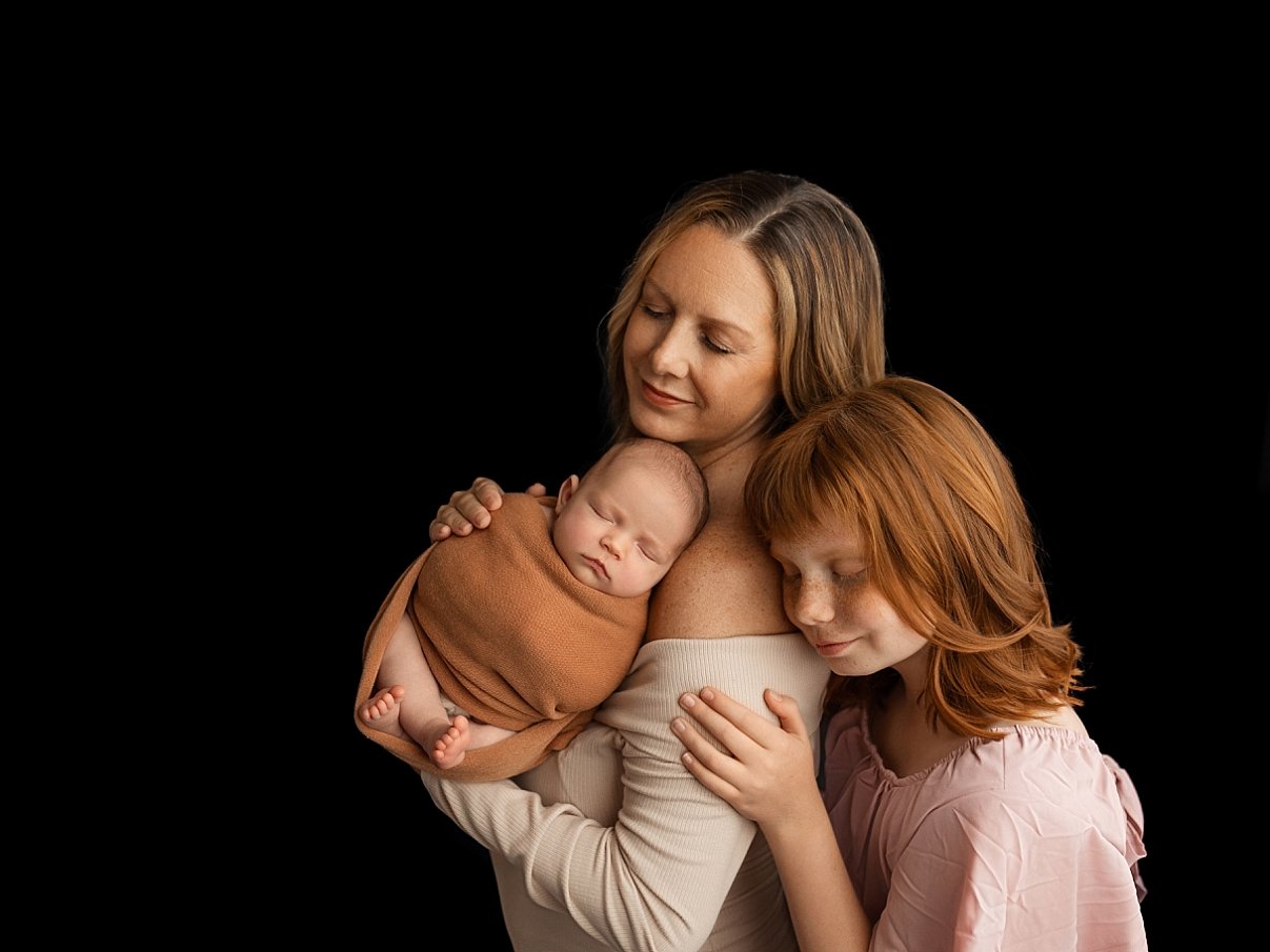 A professional fine art studio portrait by SunnyMelon Photography featuring a mother and her young daughter embracing a newborn baby. The infant is peacefully asleep, snugly wrapped in a warm terracotta-colored swaddle while resting on the mother's s