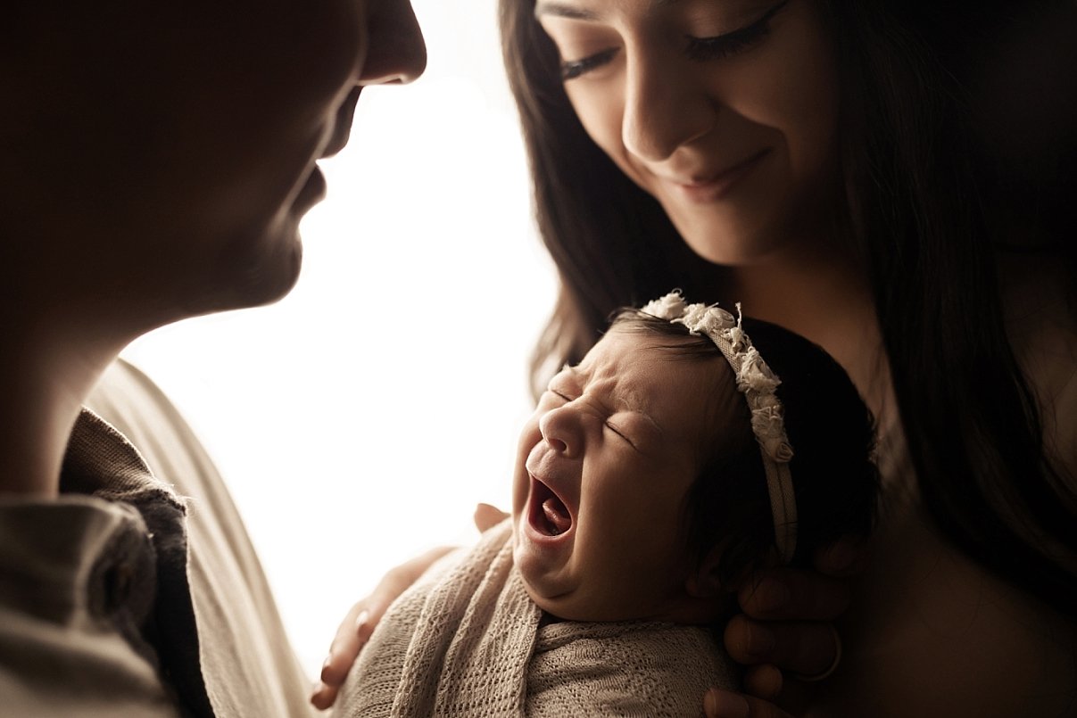 A high-contrast family photograph featuring a newborn baby held in the center, captured mid-cry or yawn with eyes squeezed shut. The infant wears a white lace floral headband and is wrapped in a neutral tan swaddle. The mother, with long dark hair, i