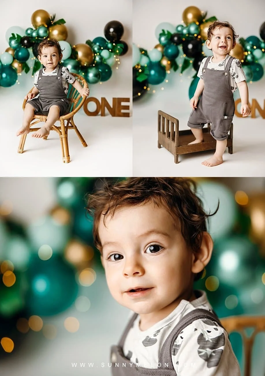 Toddler boy in grey overalls celebrating his first birthday with a green and gold balloon garland in a photography studio.