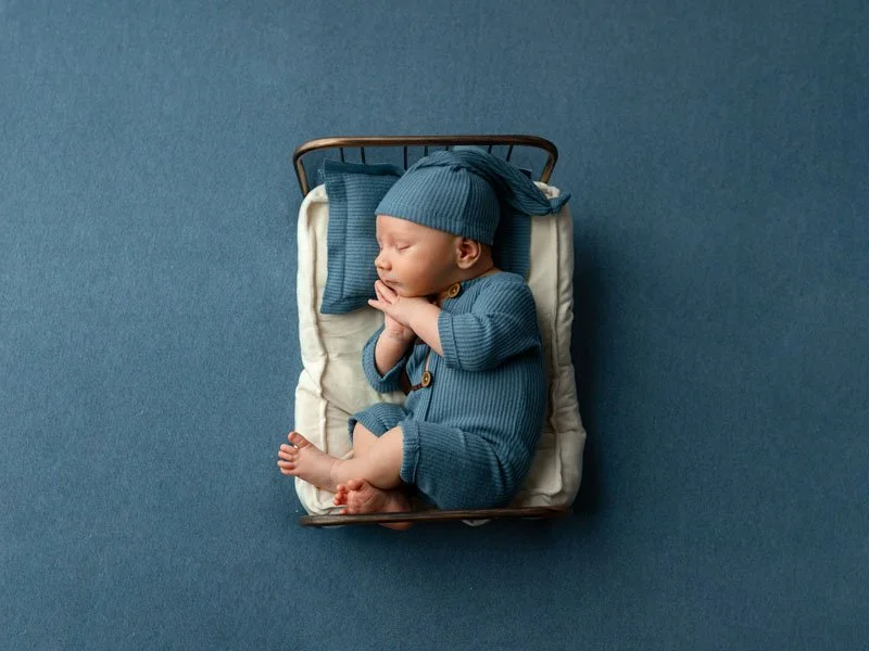 A professional overhead studio portrait of a sleeping newborn baby boy resting in a miniature vintage metal bed. The infant is dressed in a textured slate blue ribbed sleeper with wooden buttons and a matching long-tail sleepy hat. He is posed on his