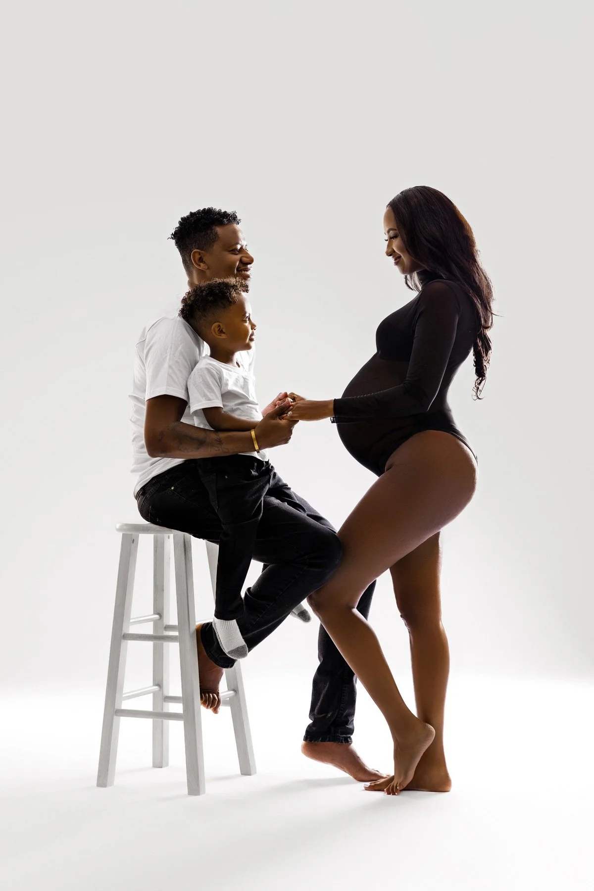 Sunnymelon Photography joyful family maternity portrait in a bright Portland studio. The pregnant mother in a black bodysuit holds hands with the father and their young son, who is sitting on a white stool.