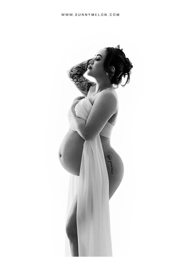 A black-and-white profile portrait of a pregnant woman with a floral arm tattoo, draped in a sheer white fabric that highlights her silhouette against a bright white background.