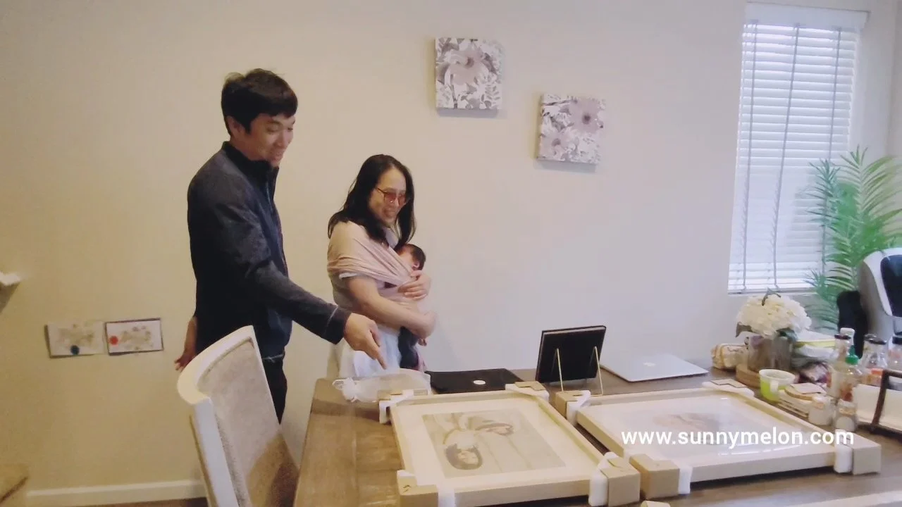 A heartwarming indoor scene where a father points toward newly delivered, professionally framed wall art resting on a dining table. The mother stands nearby, holding their newborn baby in a wrap, as they admire the large-scale prints. The image refle