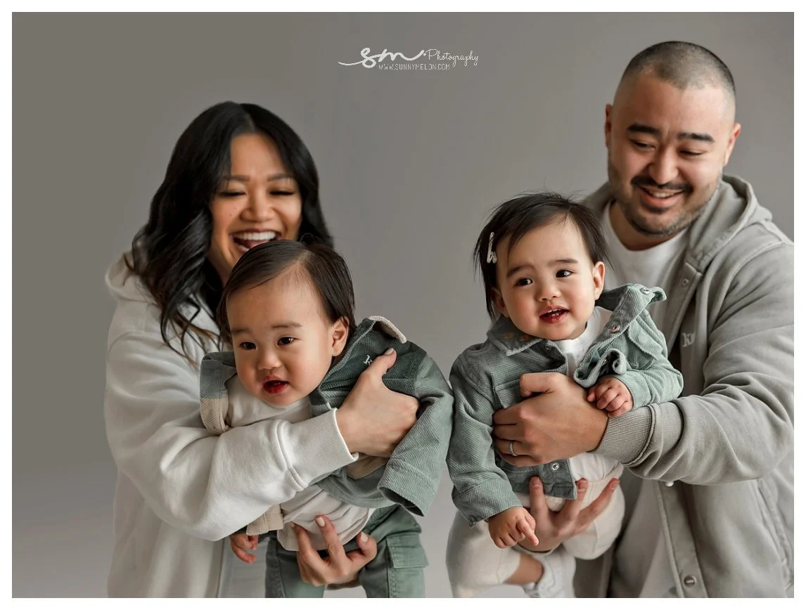 A close-up studio family portrait against a grey background; a laughing mother and smiling father hold their twin toddlers forward, who are dressed in matching sage green corduroy jackets.