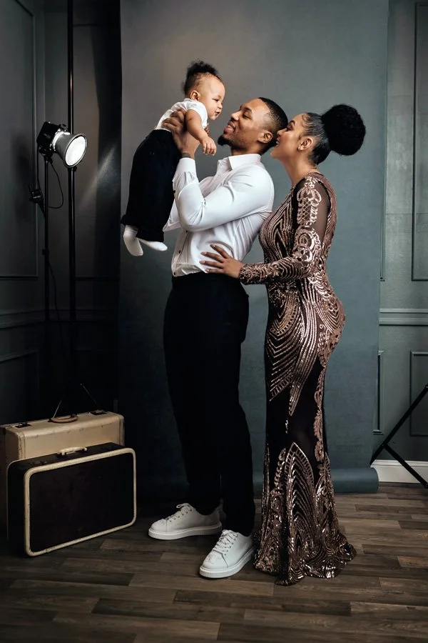 A professional studio portrait of Damian Lillard holding his young son up in the air while standing beside his partner, Kay'La. They are in formal attire against a charcoal gray backdrop with vintage suitcases on the floor.