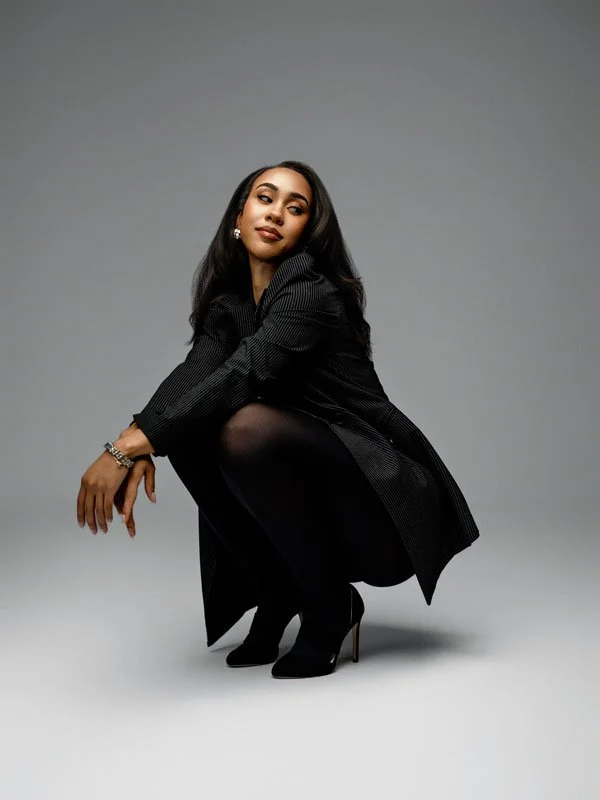 A stylish woman in a pinstripe blazer, black tights, and high heels squatting in a graceful pose against a seamless light grey studio background.
