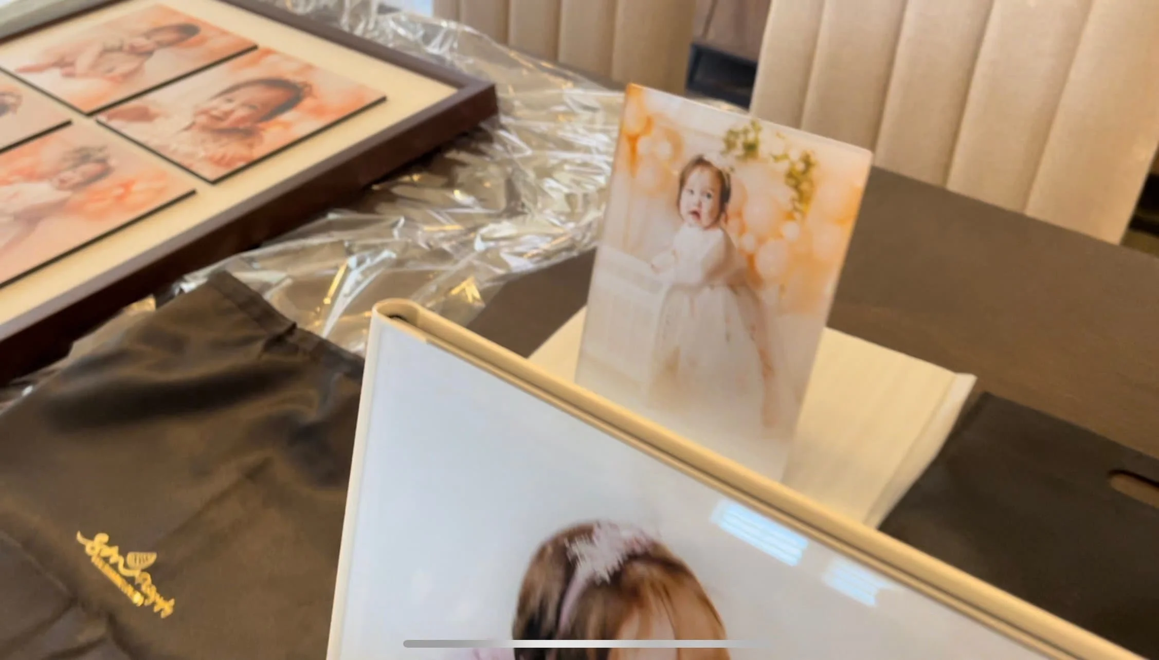 A close-up view of a professional photography product collection on a table, featuring a sleek leather-bound photo album, a standalone clear acrylic block print of a baby milestone session, and a large matted multi-aperture frame in the background.