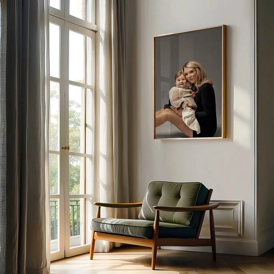 A large, elegant gold-framed portrait of a mother and daughter hanging on a white wall in a sunlit room with a modern green chair.