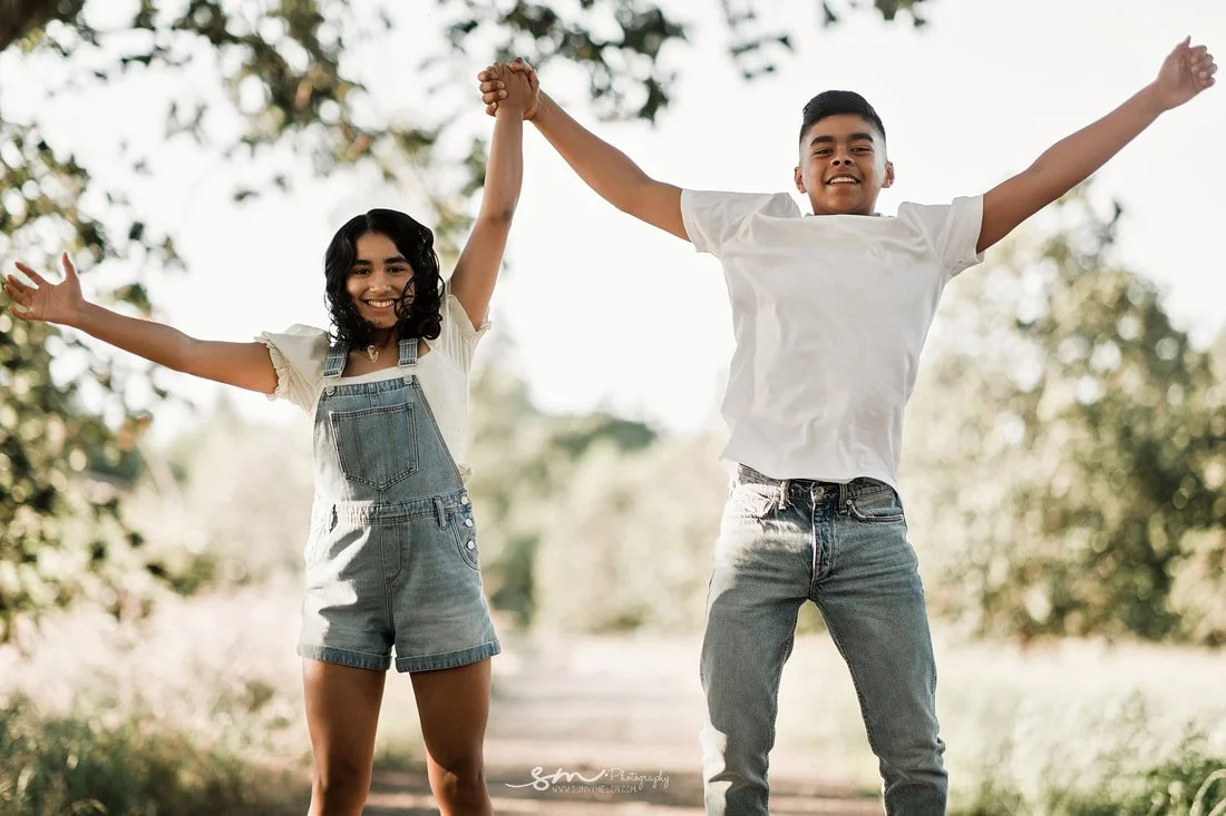Candid outdoor lifestyle portrait of teenage siblings joyfully jumping mid-air while holding hands in a bright, sunlit park setting in Portland, Oregon.