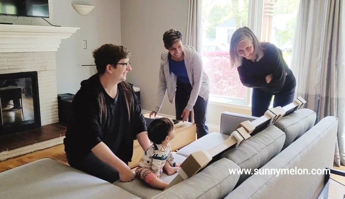 Joyful family and grandmother interacting with a baby in a living room, with framed artwork and prints from their Tualatin, Oregon outdoor family photography session being admired on the couch.