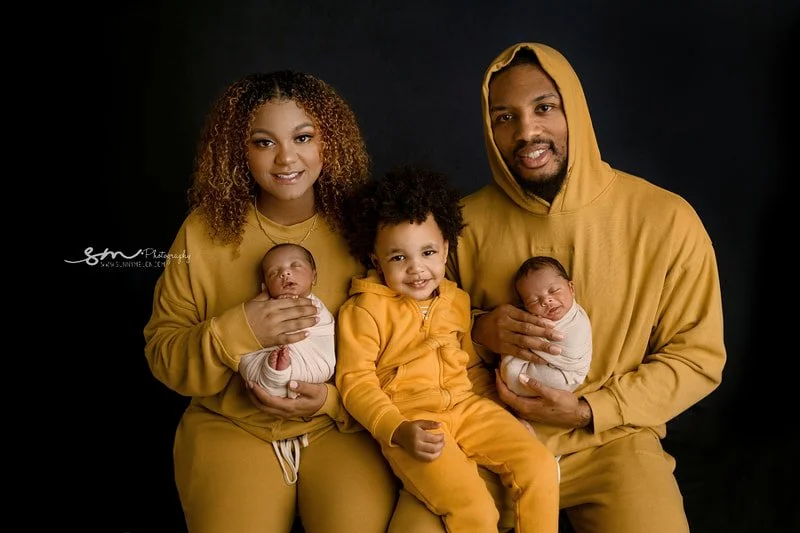 Full family portrait of Damian Lillard, Kay'La Lillard, their son, and their newborn twins (swaddled in parents' arms) all wearing matching mustard yellow tracksuits against a black background.