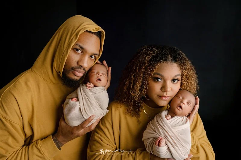 Damian Lillard and Kay'La Lillard posing for a family portrait in Portland, Oregon, holding their swaddled newborn twins while wearing matching mustard yellow hoodies against a dark background.