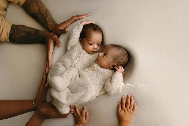 Close-up family portrait of the Lillard newborn boy/girl twins in matching knit outfits, lying face-to-face, surrounded by the loving hands of their parents and sibling during a Portland, Oregon photo session.