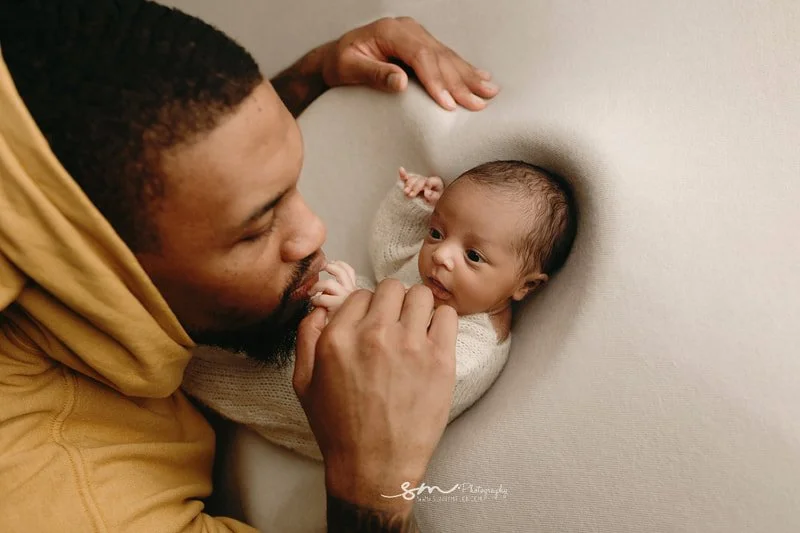 Tender portrait from the Lillard twin newborn session in Portland, Oregon, showing Damian Lillard in a yellow hoodie gently touching the face of his wide-eyed baby twin on a cream blanket.