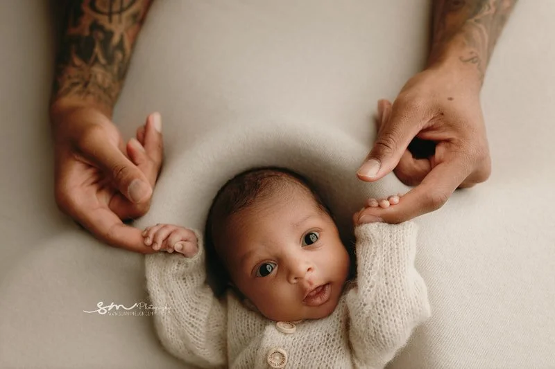 Close-up portrait of one of the Lillard newborn twins, awake and looking at the camera in a knit sweater, with a tattooed parent's hands gently holding the baby's tiny fingers during a Portland, Oregon session.