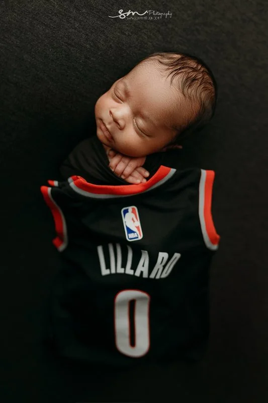 Close-up portrait of a sleeping newborn twin baby on a black background, posed next to a miniature black basketball jersey personalized with the name 'LILLARD' and number '0'.