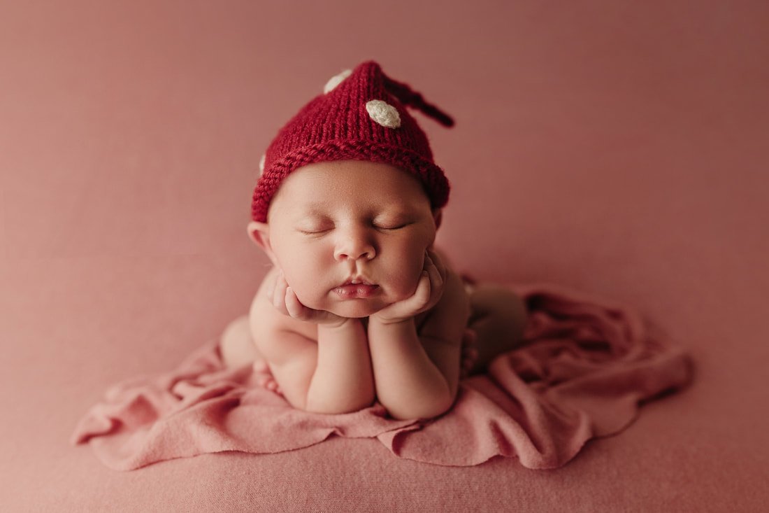 A close-up studio portrait of a sleeping newborn baby in a "chin-on-hands" pose. The infant is wearing a red knit pointed hat with white spots and is resting on a matching pink textured backdrop.