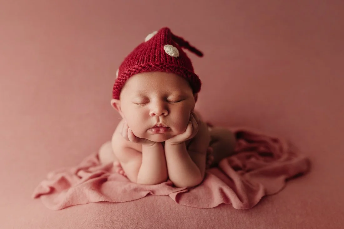 Adorable newborn baby sleeping peacefully on a pink blanket, wearing a cute red knitted hat with white polka dots, posed for a classic studio session in Portland (PDX).