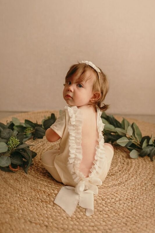 Baby in a charming ruffled romper with an open back and bow, sitting on a jute rug with eucalyptus, looking over her shoulder during a milestone session in Portland, OR.