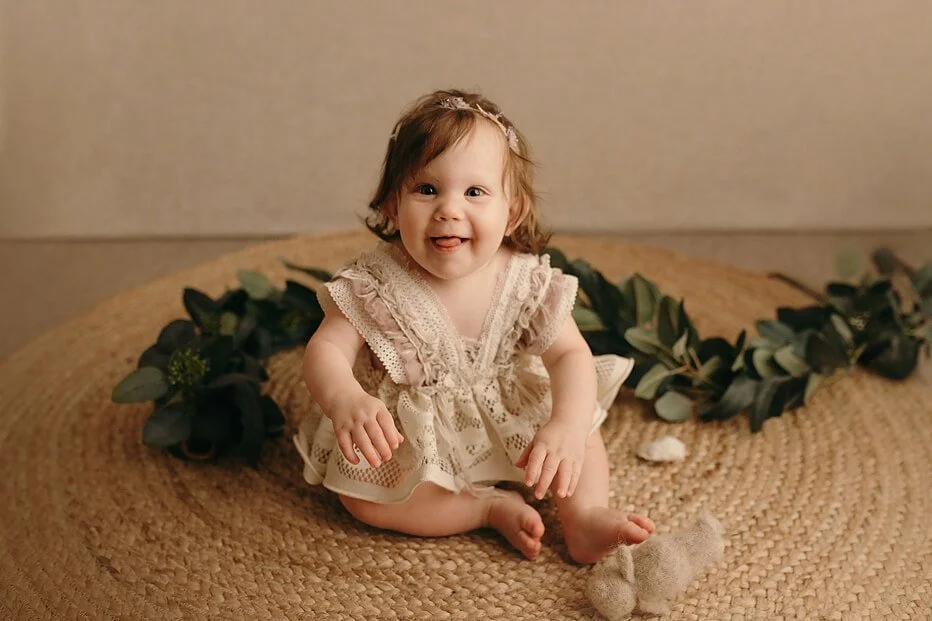 Happy baby in a lace dress and floral headband, sitting on a jute rug with eucalyptus branches, smiling at the camera for a milestone session in Portland, OR.