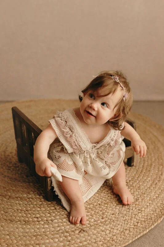 Engaged baby in a lace dress and headband, sitting in a wooden bed prop, looking up with a sweet expression during a milestone sitter session in Portland, OR.