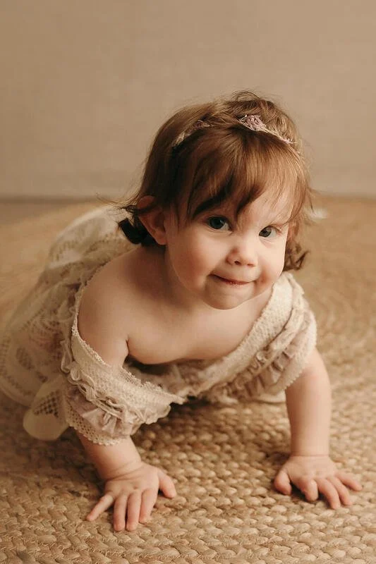 Smiling baby in a charming lace outfit, crawling on a textured jute rug, looking at the camera during a milestone sitter session in Portland, OR.