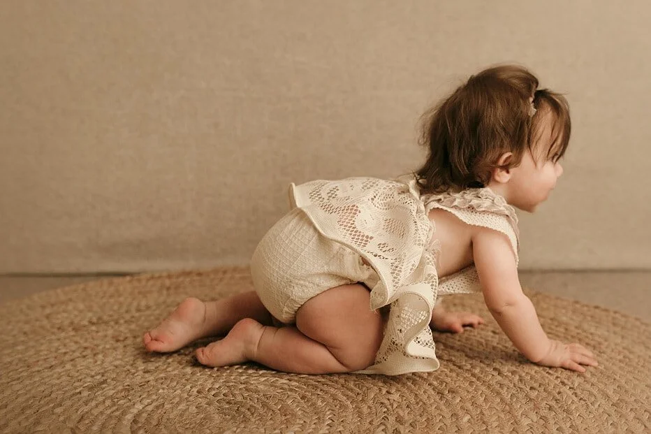 Baby in a cute lace romper crawling on a round jute rug, captured from the side during a milestone sitter session in Portland, OR.