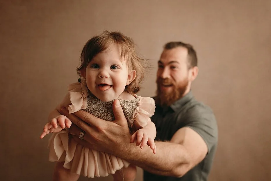 Playful baby sticking out tongue, held by a smiling dad, captured during a lively milestone sitter session in Portland, OR.