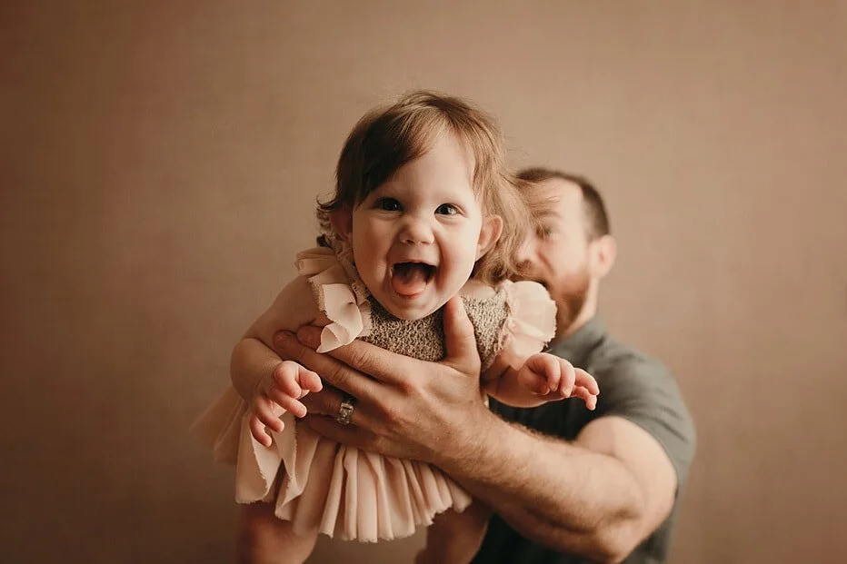 Happy baby laughing with tongue out, held securely by dad, capturing a fun milestone portrait during a sitter session in Portland, OR.